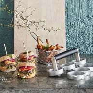 En bild visar hamburgare, pommes frites och en hamburgare-press på ett bord med en växt i bakgrunden.
