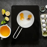 En stekpanna med ägg står på en svart skärbräda bredvid citronhalvor, en kopp dryck och en äggkartong.