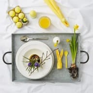En tallrik med påskägg och kvistar står på en bricka tillsammans med bestick, gula blommor och citrusfrukter.