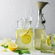 En glasburk med citronskivor och is står bredvid en flaska och ett glas med dryck på ett bord med citronfrukter och blad.