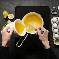 En person vispar ägg i en skål bredvid citrusfrukter, en äggkartong och kryddor på en mörk yta.