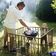En person grillar vid en rund grill medan ett litet barn tittar på från innanför ett staket.