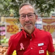 En person med glasögon och skägg står framför frukt och grönsaker i en butik.