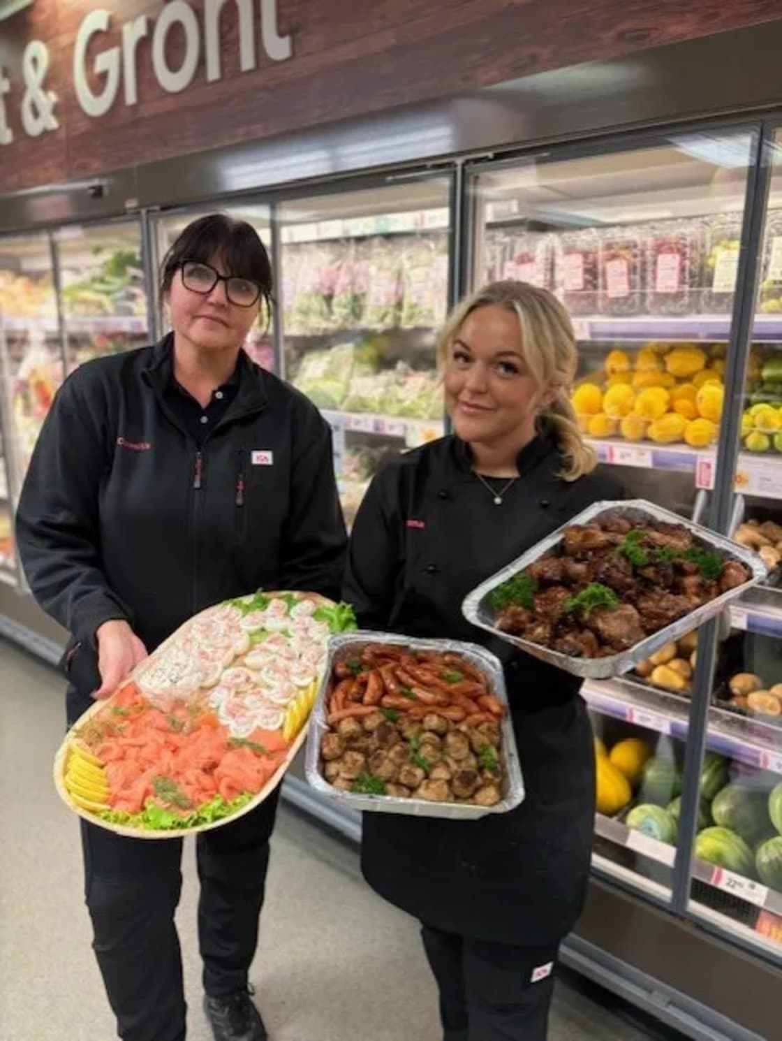 Två butikspersoner håller serveringsfat med olika pålägg, korv och grillat kött i en mataffär.
