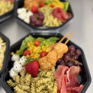 Åttakantig plastlåda med pesto-pasta, kycklingspett, lufttorkad skinka, mozzarellabollar, sallad och druvor.