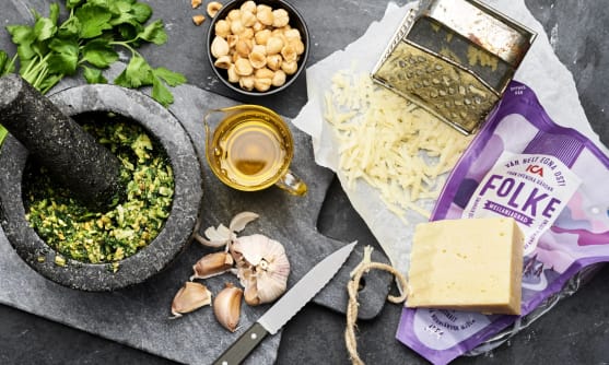 ICAs ost Folke med förpackning på ett bord tillsammans med rivjärn, riven ost och ingredienser och redskap för att göra egen pesto.
