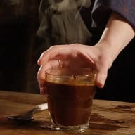 En person håller ett glas med mörk dryck över ett träbord bredvid en förpackning.