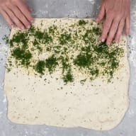 En person förbereder en deg med ost och örter på en yta.
