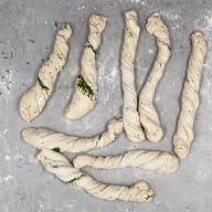 Flera avlånga degremsor ligger på en yta med några gröna örter synliga i degen.