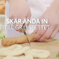 En hand med en kniv skär ett snitt i en grillkorv, som i sin tur är trädd på ett grillspett.