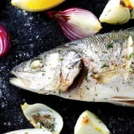 En fisk ligger på en mörk yta omgiven av bitar av lök och vitlök.
