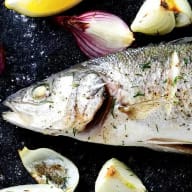 En fisk ligger på en mörk yta omgiven av bitar av lök och vitlök.