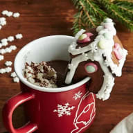 Två muggar med varm choklad och marshmallows står på ett träbord bredvid grangrenar och snöflingor.