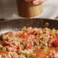 En person strör kryddor över en maträtt med köttfärs, tomater och grönsaker i en stekpanna.