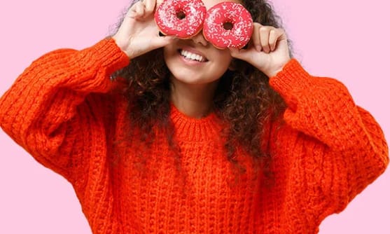 En tjej i röd stickad tröja som håller två rosa munkar framför ögonen, med en rosa bakgrund.