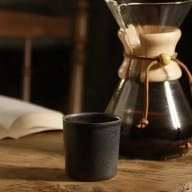 Kaffe i en pour over-kanna, intill en liten, mörkgrå keramikkopp och en uppslagen bok.