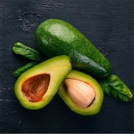 En avokado ligger bredvid två avokadohalvor med kärnor och några avokadoblad på en träyta.