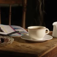 En vit kopp med rykande hett kaffe på ett träbord, omgivet av en tidning, en glasassiett och en liten mjölkkanna.