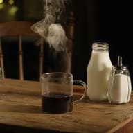 Rykande hett, svart kaffe i glasmugg på ett träbord, tillsammans med en kanna mjölk och en sockerströare.