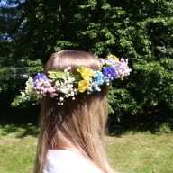 En person med långt hår bär en blomsterkrans på huvudet i en trädgård med gräsmatta och träd i bakgrunden.