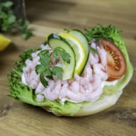 En salladsrätt med räkor, gurka, tomat och persilja serveras på ett salladsblad på ett träbord.