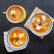 Tre skålar med soppa innehållande tärningar av tofu och grönsaker står på en mörk yta.