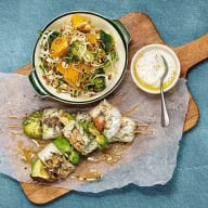 En tallrik med sallad och nudlar står bredvid en skål med sås och grillspett på en skärbräda.