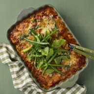 En ugnsform med zucchini melanzane står på ett underlag bredvid en tygbit och en serveringssked.