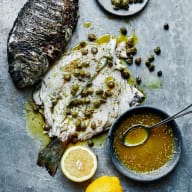 En tillagad fisk ligger på en tallrik med kapris och citron bredvid en skål med sås.